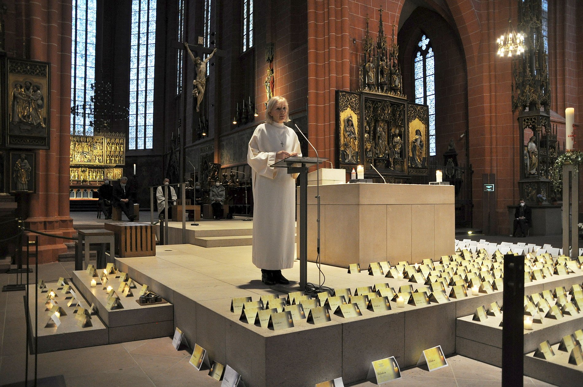 Eine Frau steht im liturgischen Gewand in einer Kirche. Ringsherum stehen unzählige Gedenkkarten für Corona-Verstorbene.